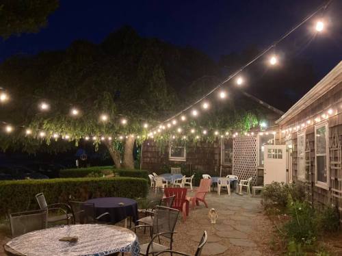 A cozy outdoor patio at night, adorned with string lights, featuring tables and chairs surrounded by greenery.