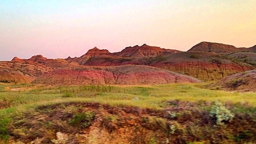 Colorful, layered rock formations rise against a pastel sky, surrounded by grassy plains in a serene landscape.