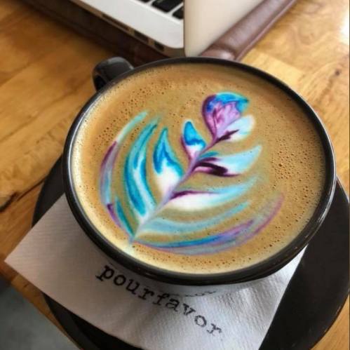 A beautifully crafted latte with colorful floral latte art, served in a black cup on a wooden table.