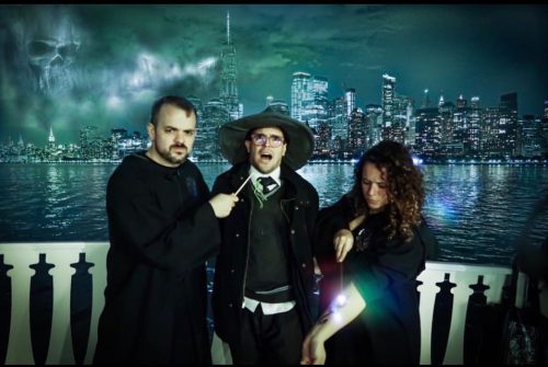 Three people in dark cloaks pose against a city skyline at night, with one holding a glowing object and a spooky backdrop.