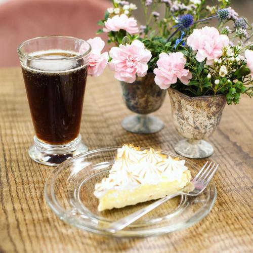 A glass of dark beverage beside a slice of meringue pie on a plate, with a small vase of pink flowers in the background.