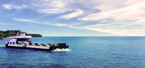 A ferry carrying cars navigates through calm blue waters under a partly cloudy sky.