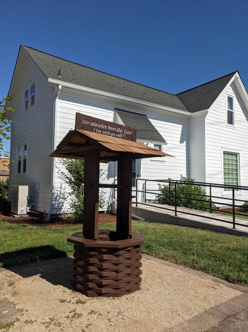 A wooden well with a sign reading "Wir wünschen Ihnen alles Gute!" in front of a white house on a sunny day.