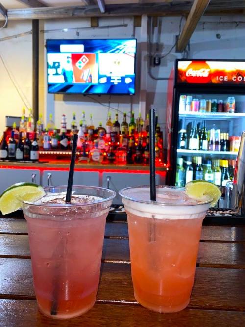 Two colorful cocktails with lime slices sit on a wooden bar, with a well-stocked back bar and a TV in the background.