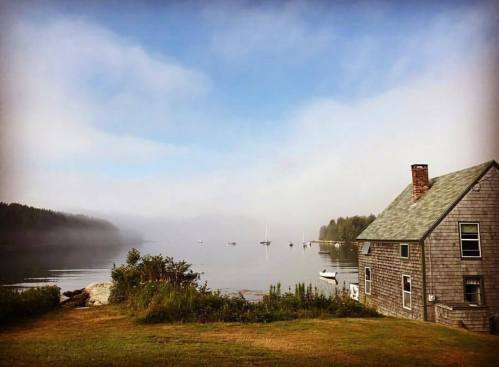 A foggy morning by the water, featuring a rustic house and boats anchored in a serene bay.