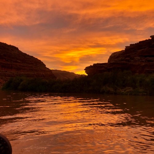 A serene river scene at sunset, with vibrant orange and purple skies reflecting on the water, surrounded by rocky cliffs.
