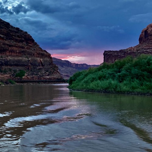 A serene river flows between rocky cliffs under a dramatic sky at sunset, with lush greenery along the banks.