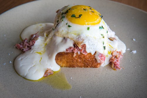 A slice of toasted bread topped with creamy sauce, shredded meat, and a sunny-side-up egg, garnished with herbs.