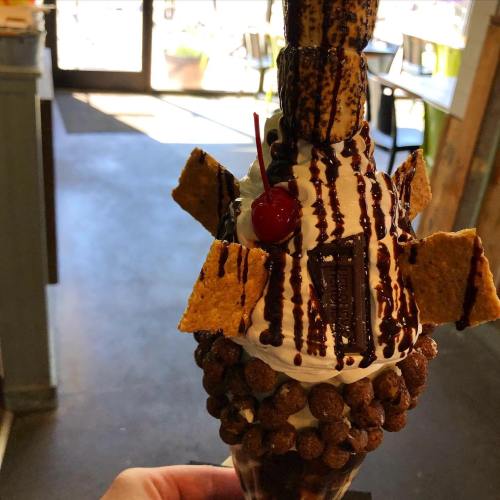 A hand holding a tall ice cream cone topped with chocolate drizzle, a cherry, and graham cracker pieces.