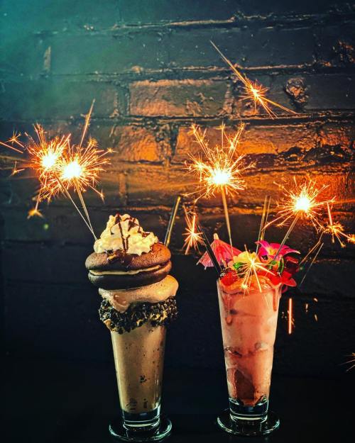 Two festive milkshakes topped with whipped cream and cookies, adorned with sparklers against a dark brick background.