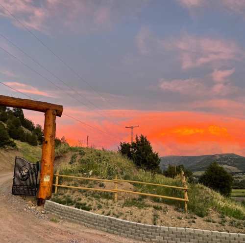 A wooden gate at sunset, with vibrant orange and pink skies above rolling hills and greenery.
