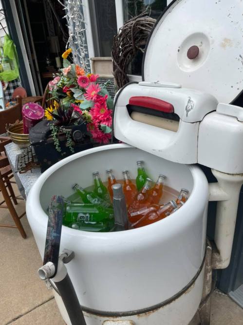 A vintage white cooler filled with colorful soda bottles, surrounded by flowers and outdoor furniture.
