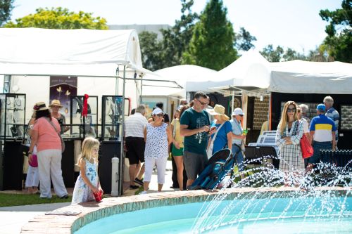 A busy outdoor market scene with people browsing tents and a fountain in the foreground. A child stands nearby.