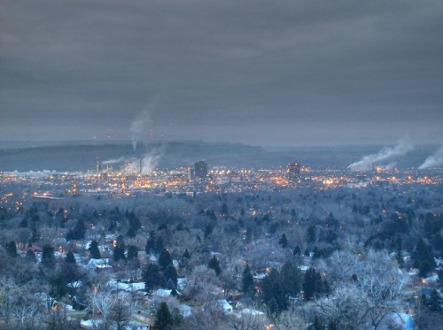 A city skyline at dusk, with industrial buildings and smokestacks emitting smoke, surrounded by trees.