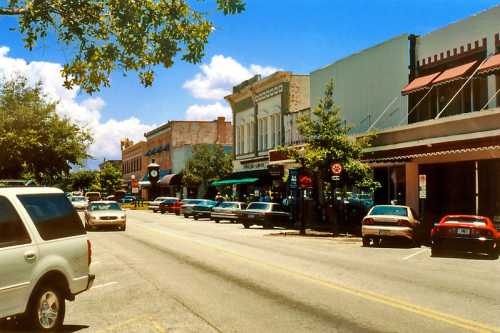 A sunny street scene featuring shops, parked cars, and trees in a small town. Bright blue sky with a few clouds.