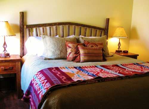 Cozy bedroom with a wooden bed frame, decorative pillows, and a colorful blanket, illuminated by two bedside lamps.