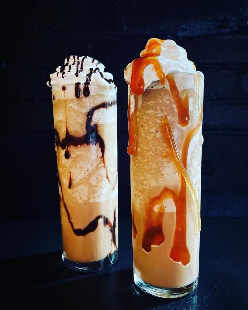 Two tall glasses of creamy beverages topped with whipped cream, chocolate drizzle, and caramel sauce against a dark background.