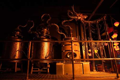 Industrial distillation equipment with metallic tanks and intricate piping, illuminated by warm, ambient lighting.