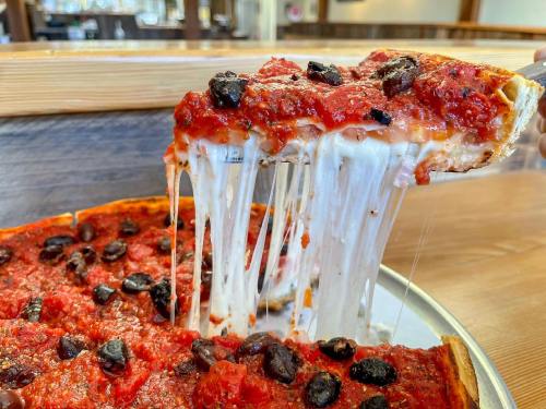 A slice of cheesy pizza with a thick tomato sauce and black olives being lifted from a pan.