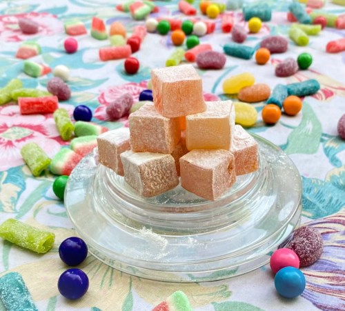 A glass dish holds colorful cubes of candy, surrounded by various sweets on a floral-patterned tablecloth.