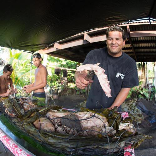 A man holds a piece of meat over a table filled with food, while two others prepare in the background.