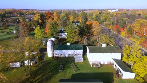 Aerial view of a farm surrounded by colorful autumn trees, featuring buildings and a silo on a sunny day.