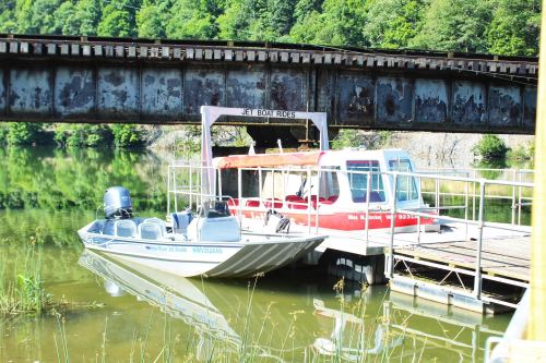 Two boats docked at a pier under a rusted railway bridge, surrounded by calm water and greenery.