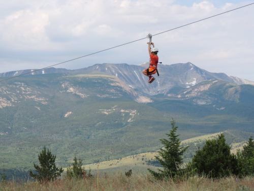 A person in an orange harness zip-lining over a mountainous landscape with green trees and a cloudy sky.