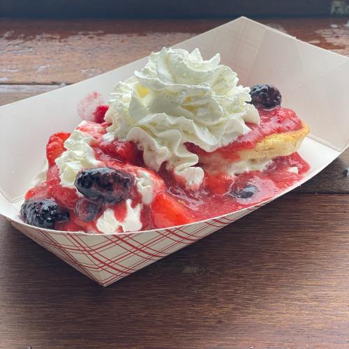 A dessert in a paper tray featuring a biscuit topped with strawberries, blackberries, whipped cream, and strawberry sauce.