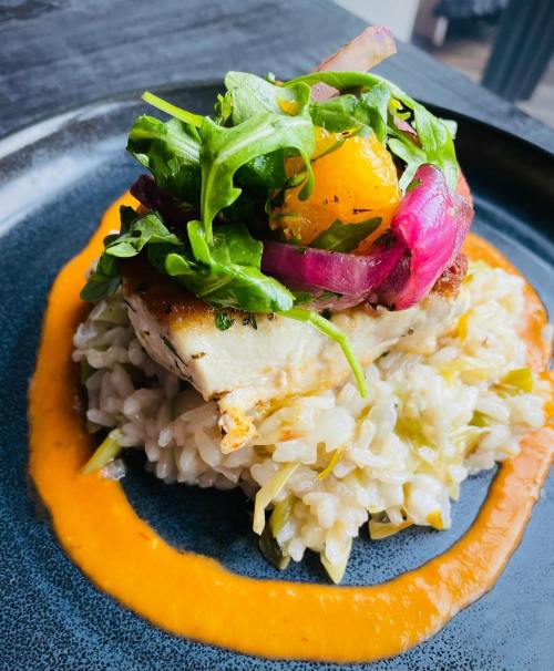 A plate of risotto topped with grilled fish, arugula, orange segments, and pickled onions, drizzled with sauce.