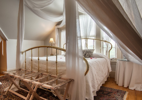 Cozy bedroom with a canopy bed, soft lighting, and elegant decor, featuring a bench and natural light from windows.