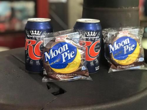 Three cans of RC soda next to two packages of chocolate Moon Pies on a black surface.