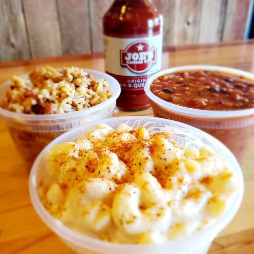 Three containers of food: macaroni and cheese, rice, and beans, with a bottle of barbecue sauce in the background.