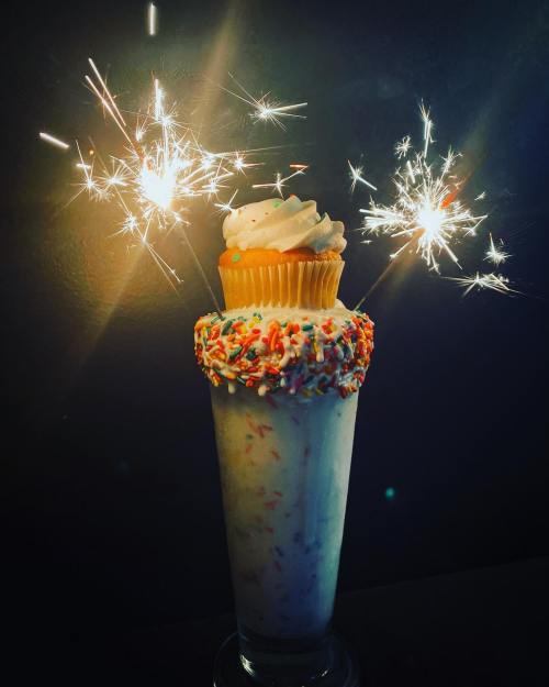 A tall milkshake topped with a cupcake and colorful sprinkles, with sparklers adding a festive touch.