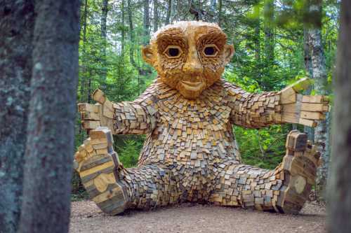 A large wooden troll sculpture sits in a forest, surrounded by trees, with arms outstretched and a friendly expression.