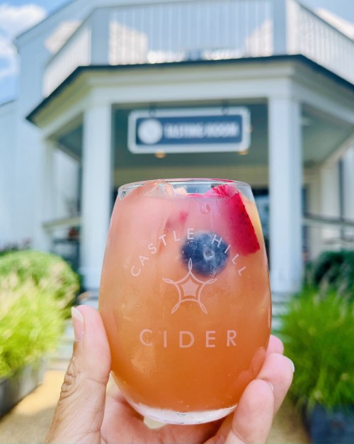 A hand holding a glass of cider with berries, in front of a tasting room building.