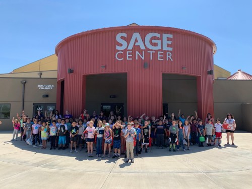 A large group of children and adults gather outside the Sage Center, smiling and waving at the camera.