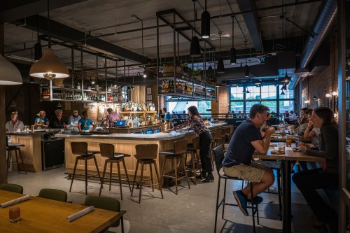 A modern bar and restaurant interior with patrons seated at tables and the bar, featuring warm lighting and a lively atmosphere.