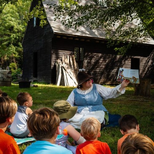 A storyteller in period costume reads to a group of children outdoors near a rustic wooden building.