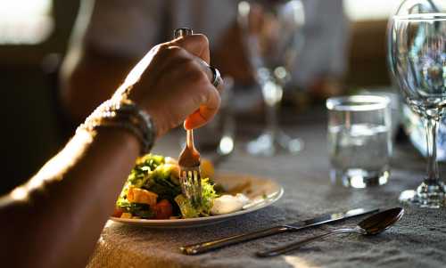 A hand holding a fork reaches for a salad on a plate, with glasses and a softly lit table in the background.