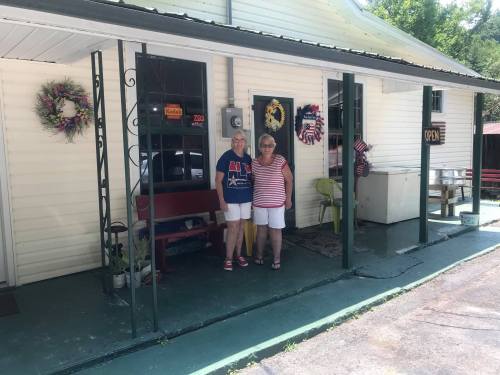 Two women stand on a porch in front of a white building, smiling, with decorative wreaths and an "Open" sign visible.