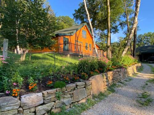 A charming orange cabin surrounded by lush greenery and colorful flowers, with a gravel path leading up to it.
