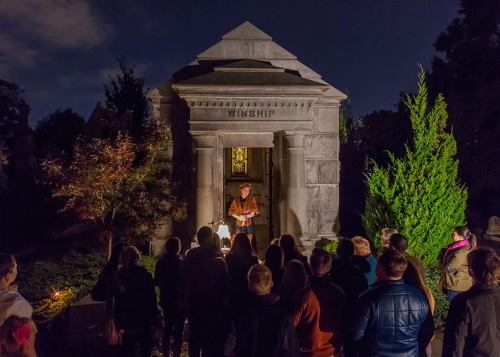 A group of people gathers at night near a stone mausoleum, listening to a speaker in front of a stained glass window.