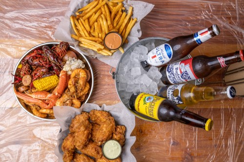 A spread of seafood, fries, and fried chicken with various beers on ice, served on a wooden table.
