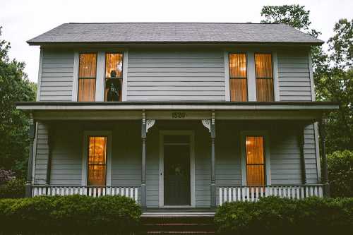 A two-story gray house with lit windows, surrounded by greenery, and a figure visible in the upper window.