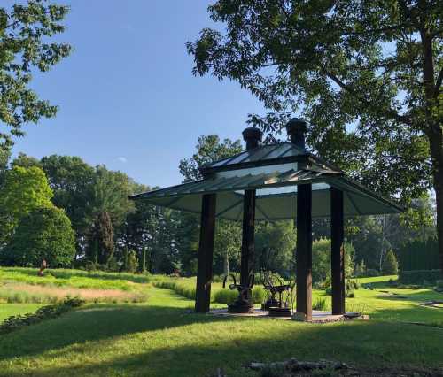 A gazebo with a metal roof stands in a lush green garden under a clear blue sky.