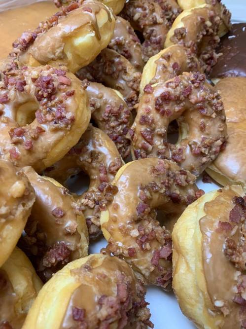 A close-up of glazed donuts topped with chopped bacon, arranged in a pile with a chocolate donut in the background.