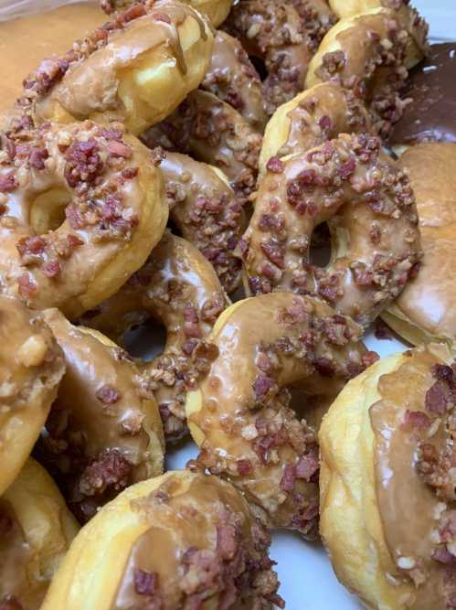 A close-up of glazed donuts topped with chopped bacon, arranged in a pile with a chocolate donut in the background.