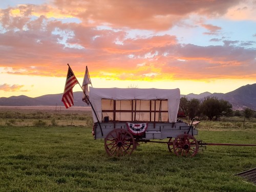 A covered wagon with flags stands in a grassy field against a vibrant sunset and mountains in the background.