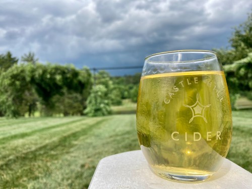 A glass of cider sits on a table, with a lush green landscape and cloudy sky in the background.